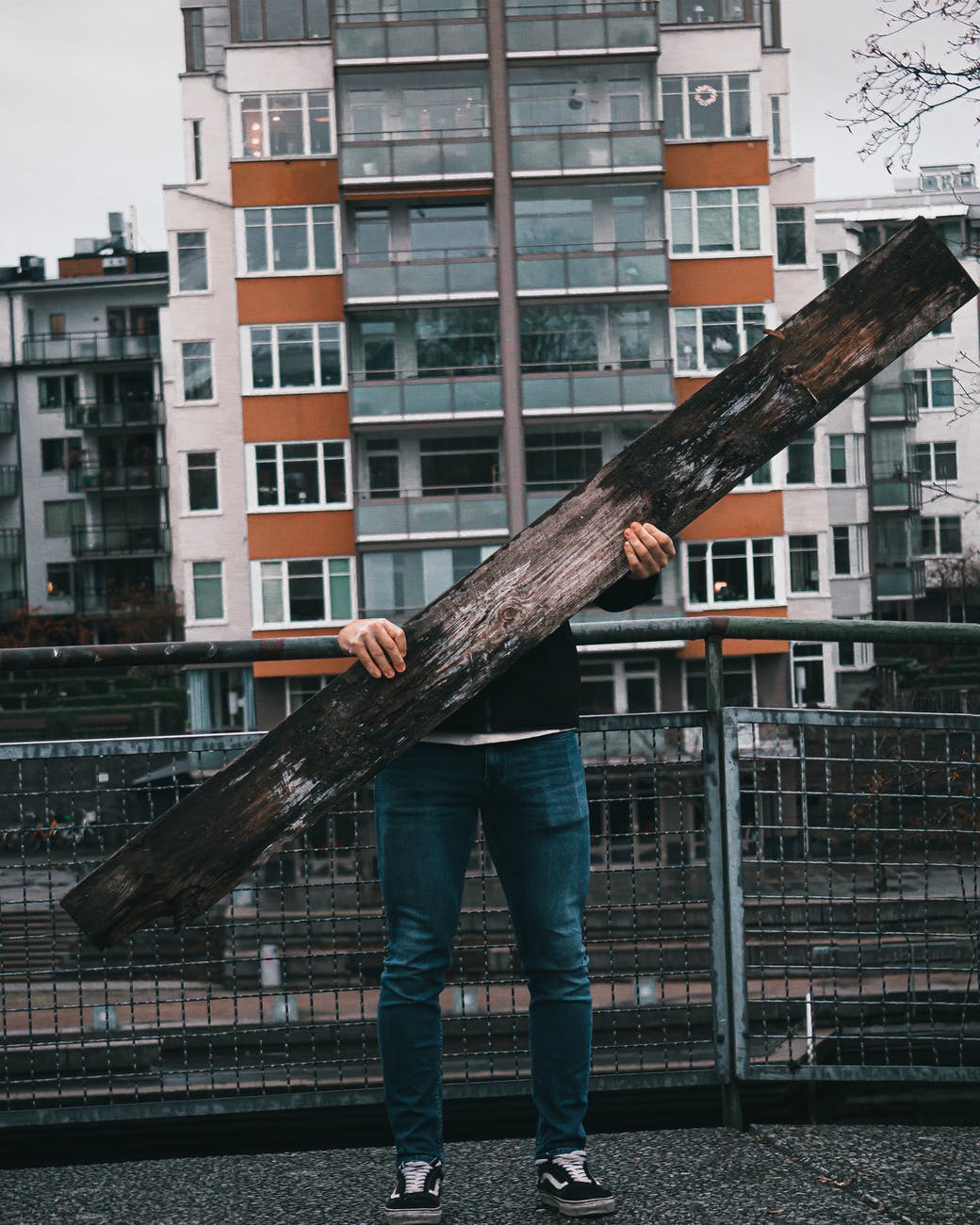 A person in blue denim jeans standing holding brown wooden log. Person is not visible above the board, and you see the building in the background